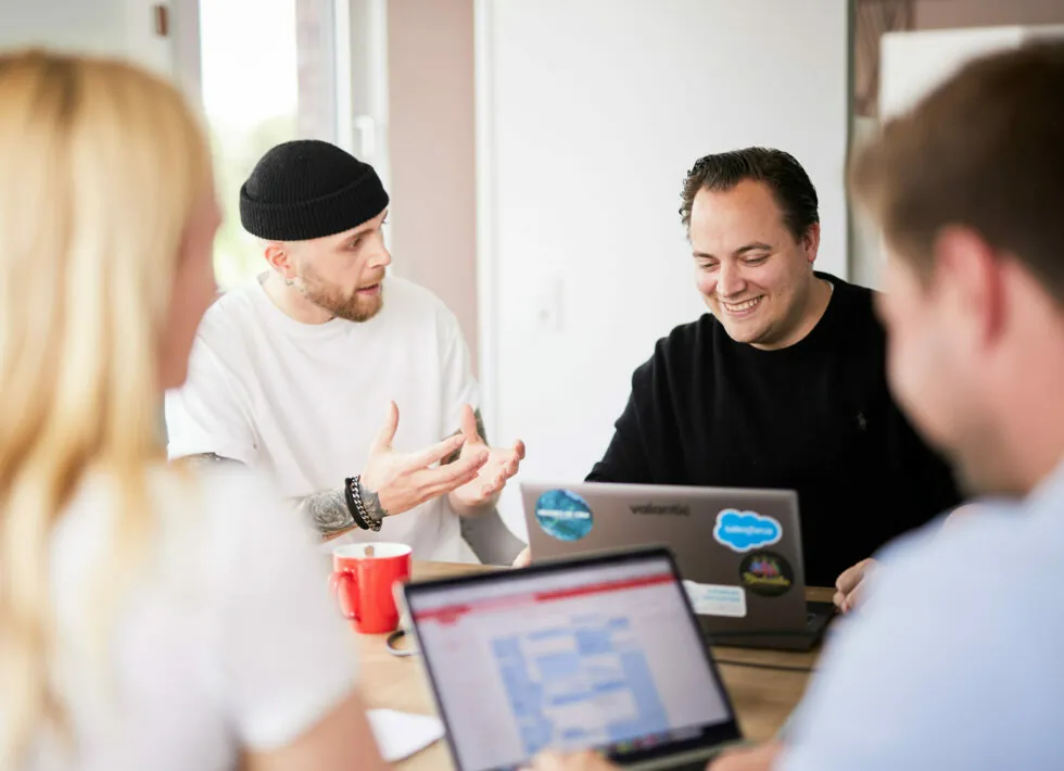 Four individuals are sitting around a table with laptops, having a discussion. Two men in the center are engaged in conversation, one gesturing with his hands, the other smiling.