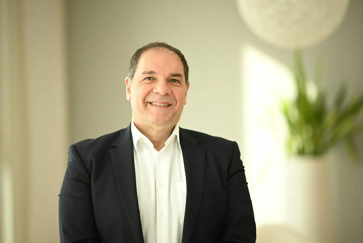 A middle-aged man in a suit jacket and white shirt smiles at the camera. A potted plant and soft lighting are in the background.