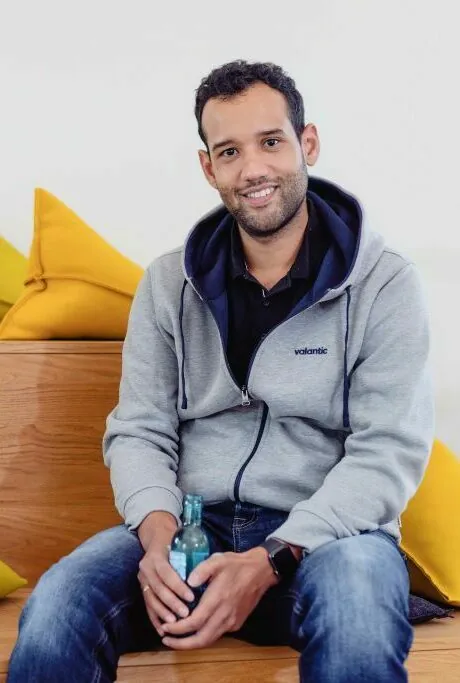 A man wearing a gray hoodie and jeans sits on wooden steps with yellow cushions in the background, holding a bottle and smiling at the camera.