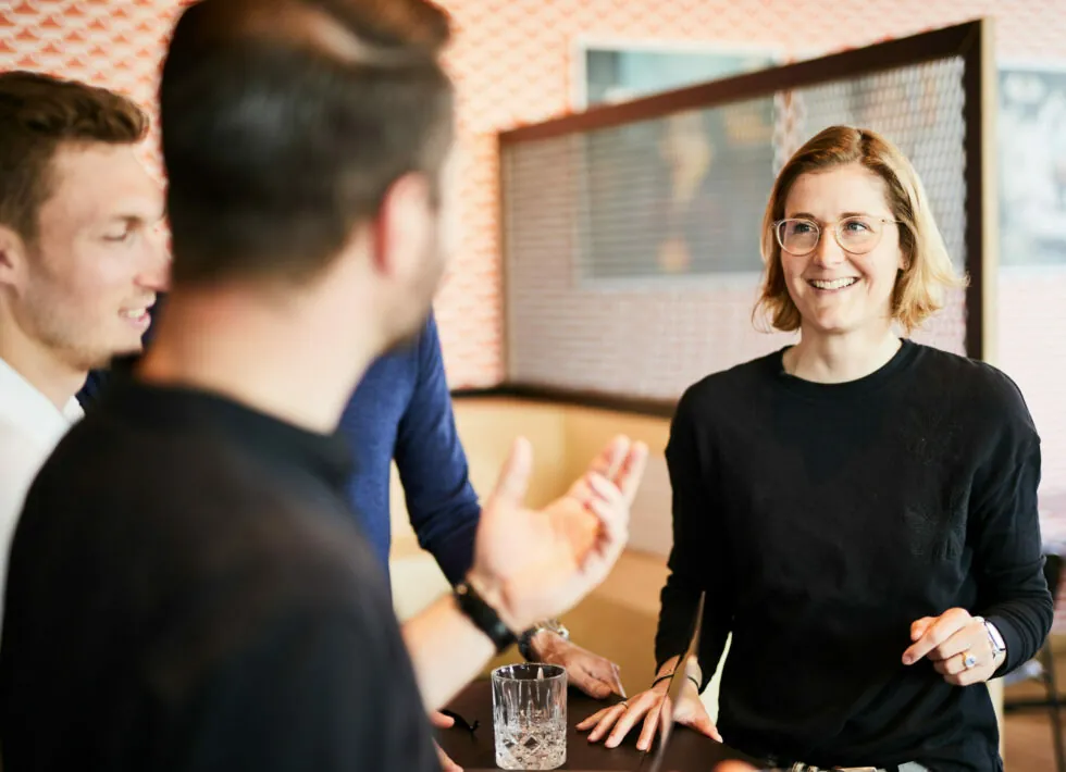 Three people are engaged in a conversation at a casual indoor setting. A woman with glasses is smiling, while the two men, one blurred in the foreground and one on the left, listen and speak.