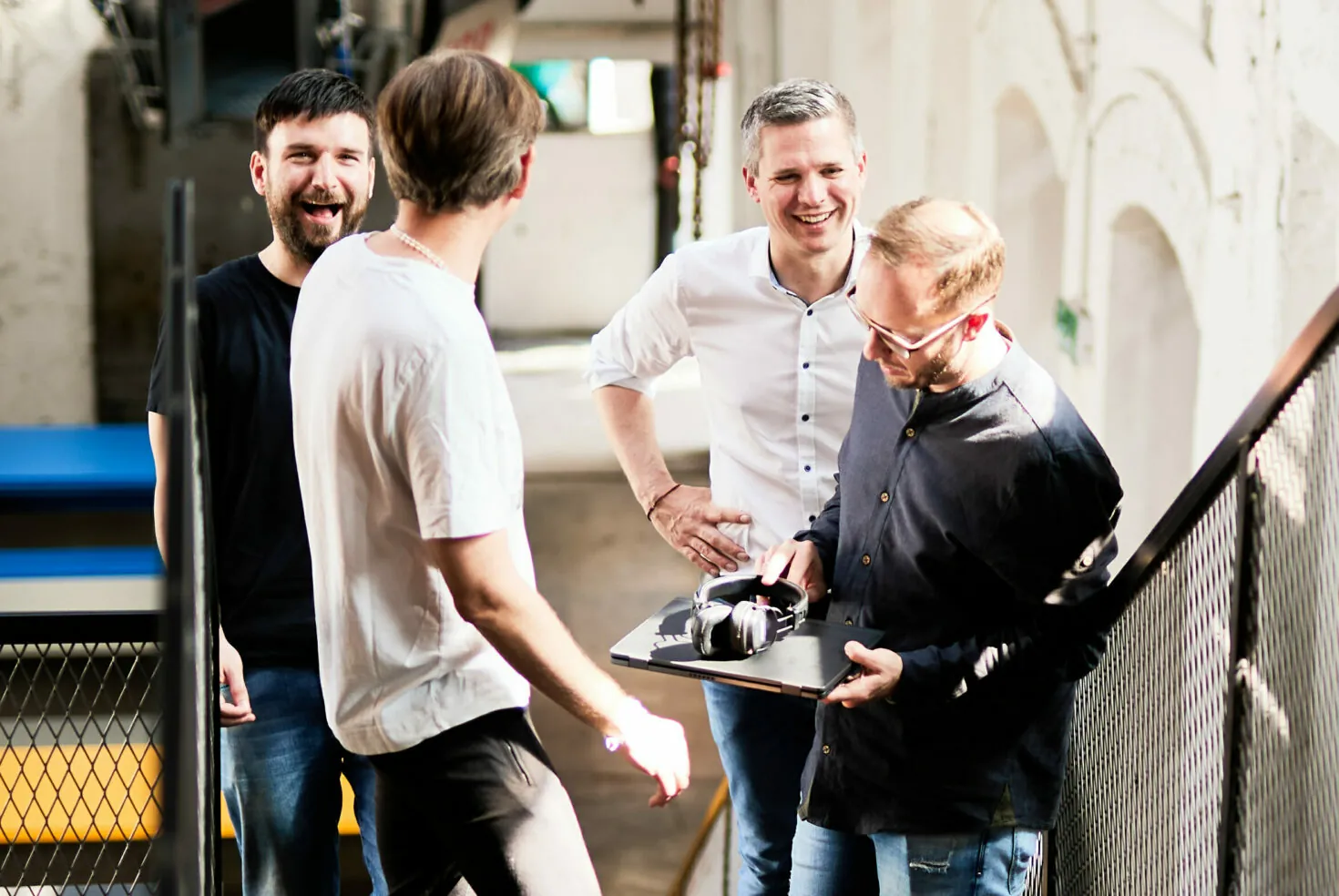 Four men are gathered on a stairwell, engaged in conversation and smiling. One man holds a clipboard and headphones.