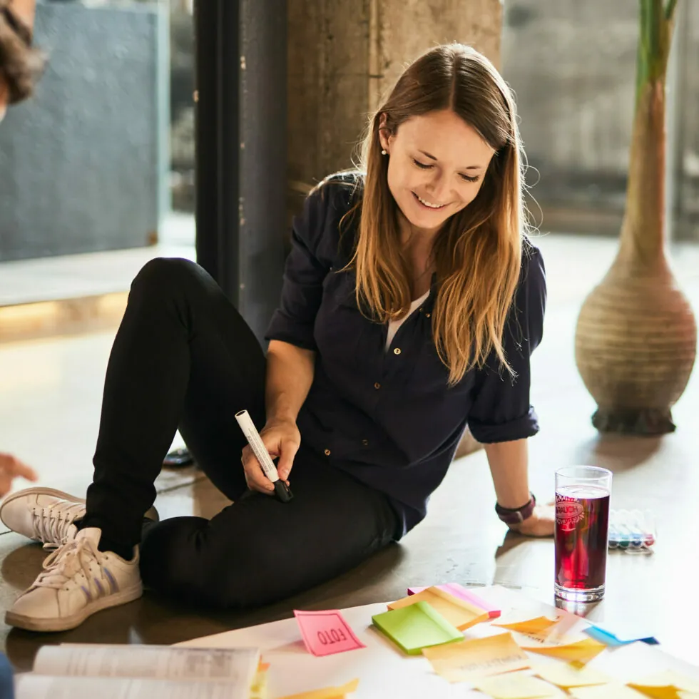 Woman sits on floor, smiling, and writes on colorful sticky notes spread out in front of her. She's holding a marker with a glass of red drink beside her.