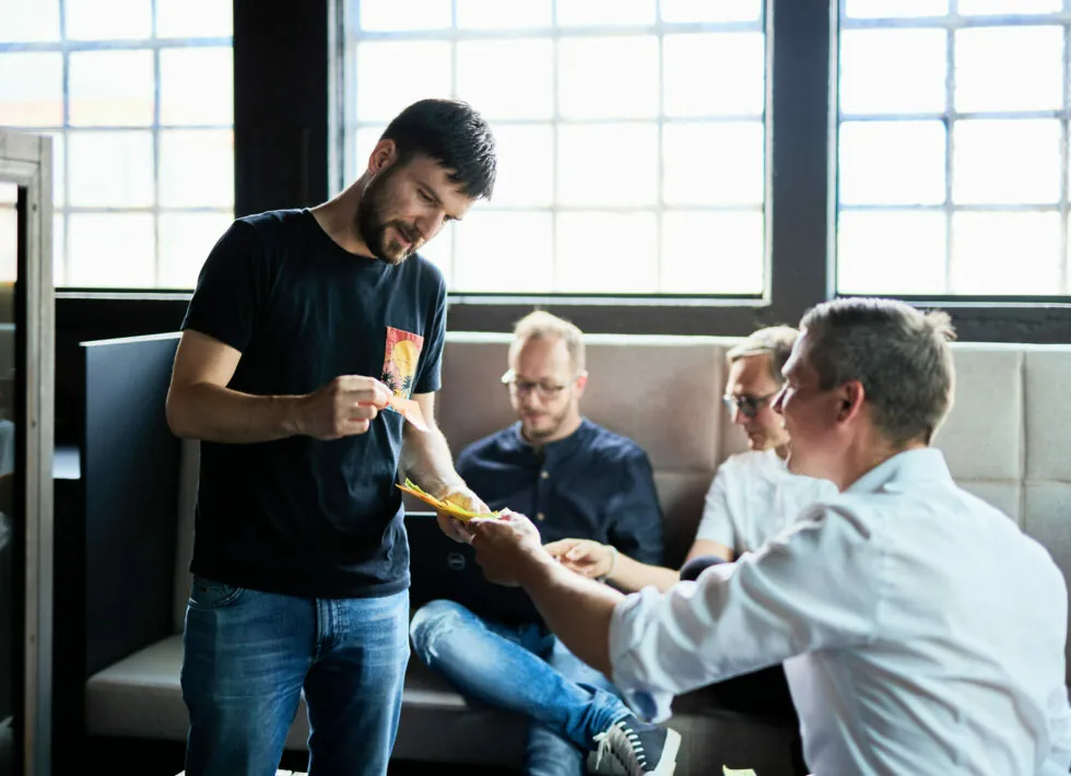 A man hands food to another seated man in an office setting, with two other individuals sitting and working on laptops in the background.