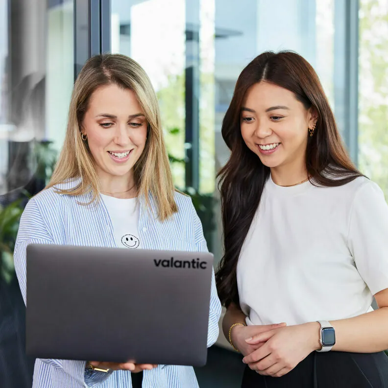 Two women standing and smiling while working on a laptop computer with the brand name "valantic" visible on the device.