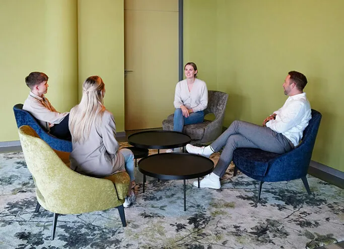 Four people are seated in a casual meeting space with green walls, upholstered chairs, and round tables, engaged in conversation.