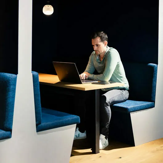 A man sits in a booth working on a laptop, with dark blue upholstery and a small overhead light.