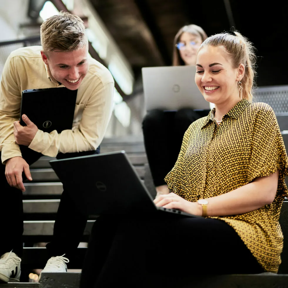 Three young adults are seated on stairs and using laptops. One person standing holds a laptop while interacting with a person sitting, who is smiling. The third person in the background is also using a laptop.