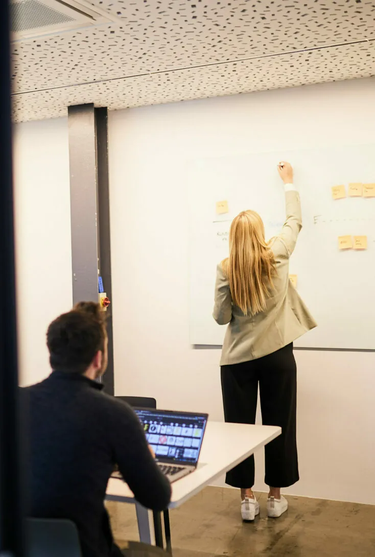 A woman stands writing on a whiteboard with sticky notes while a seated man works on a laptop, facing her in a modern office setting.
