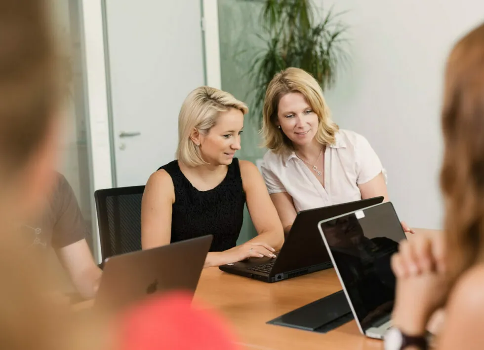 Two women sit at a meeting table while looking at a laptop together. Other participants with laptops are also present, partially visible. A plant and a glass door are in the background.