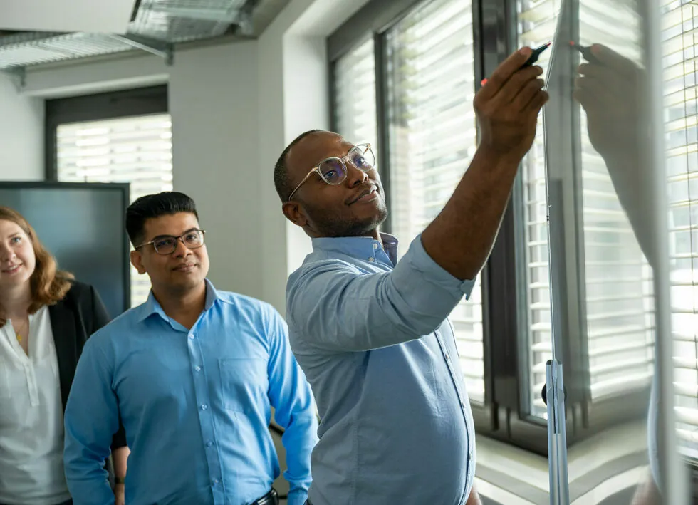 A man writes on a whiteboard while two colleagues watch attentively in a modern office setting with large windows.