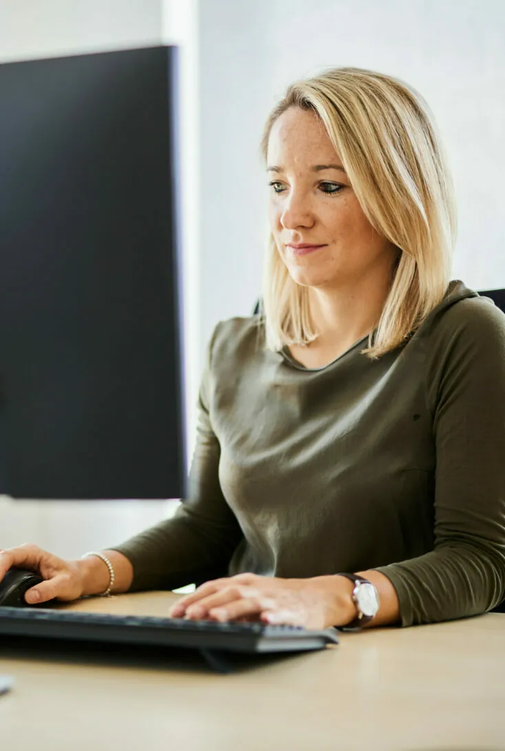 A person with shoulder-length blonde hair, wearing an olive-green top, sits at a desk and types on a keyboard while looking at a monitor.