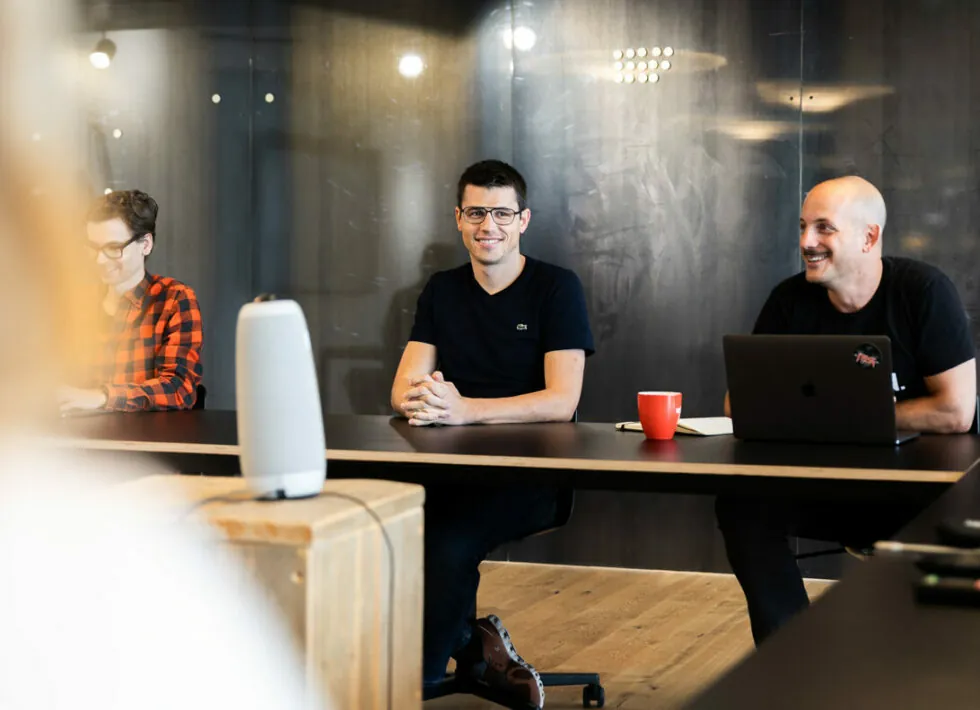 Three people are sitting at a table in an office setting, one with a laptop, and a speaker is positioned on the table in the foreground.