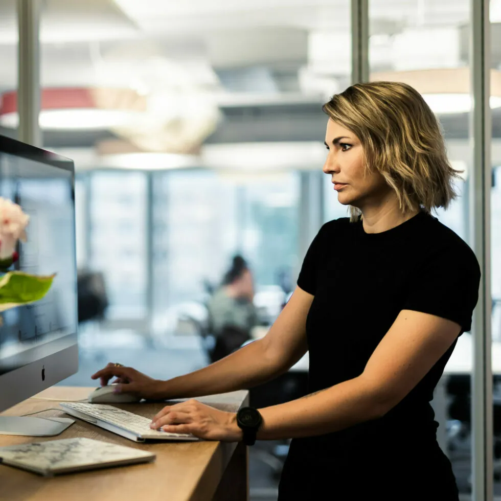 A woman is standing at a desk in an office, typing on a keyboard and looking at a computer monitor. Office environment in the background.