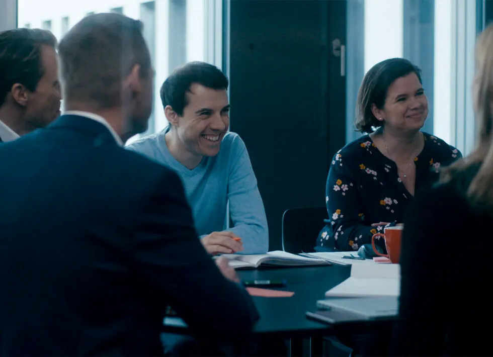A group of five people sitting around a table, engaged in discussion. One man is smiling and another woman appears attentive. Papers, notebooks, and pens are on the table.