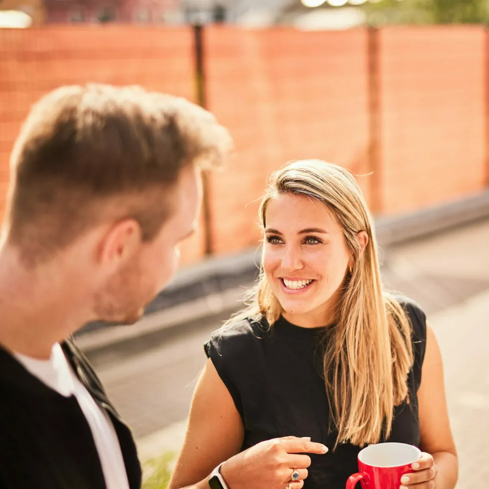 Two people are outdoors, engaged in conversation. One person is holding a red mug and smiling at the other. There is an orange fence in the background.