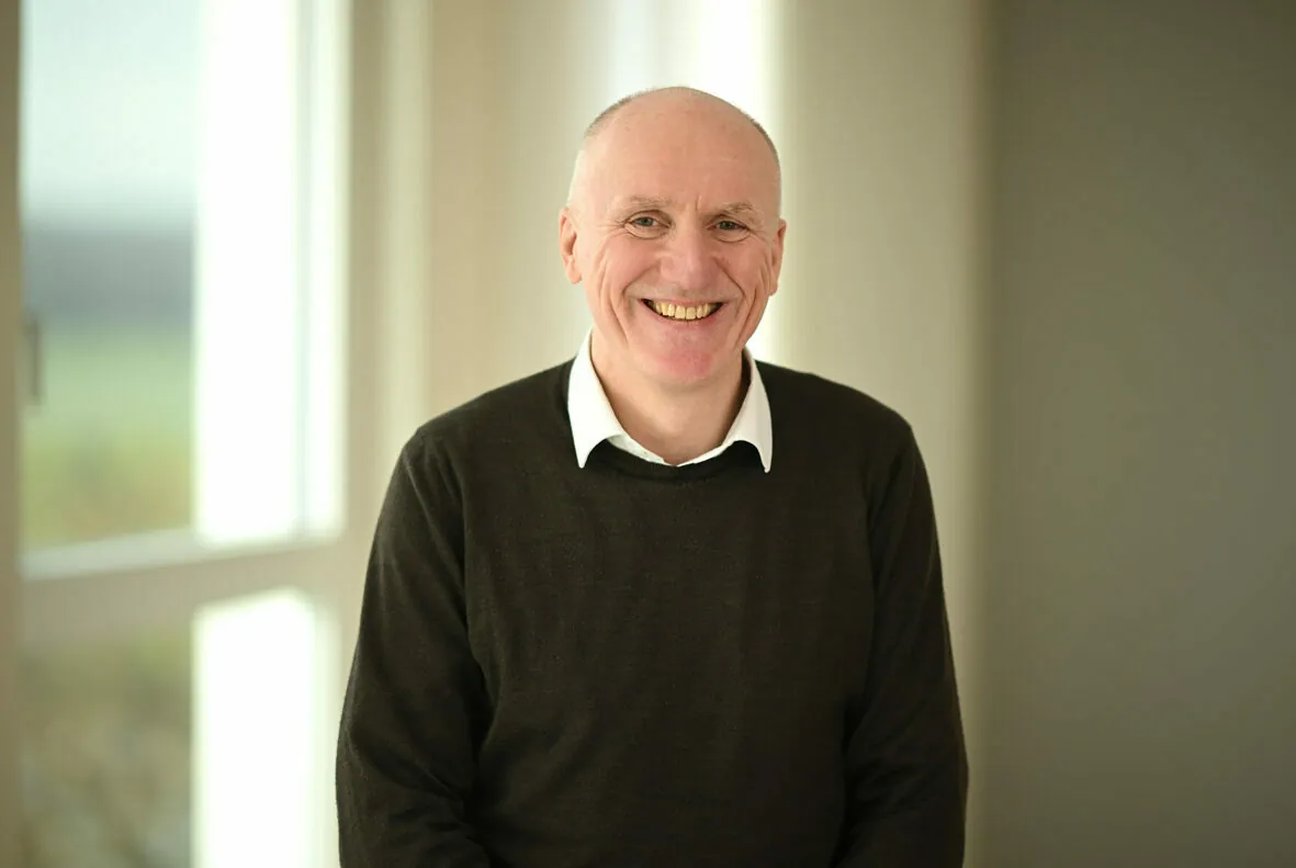 A smiling bald man in a dark sweater and white collared shirt stands in a bright room with large windows in the background.
