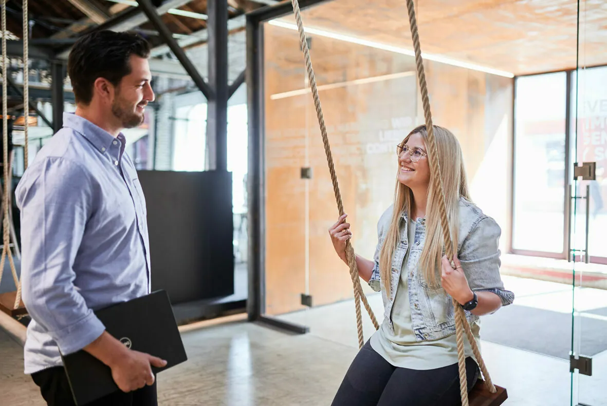 A man holding a laptop smiles and talks to a woman sitting on a swing in a modern office space.