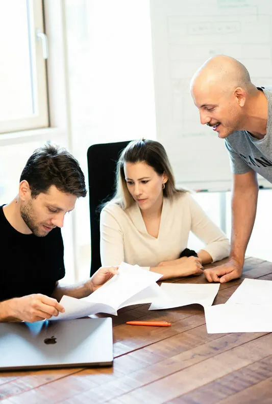 Three people collaborate around a table with documents and a laptop. One person is seated with two others standing, looking at the papers. A whiteboard in the background contains text and diagrams.