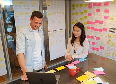 Two people are standing near a table with colorful sticky notes, looking at a laptop. Behind them, a wall is covered with additional sticky notes and planning documents.