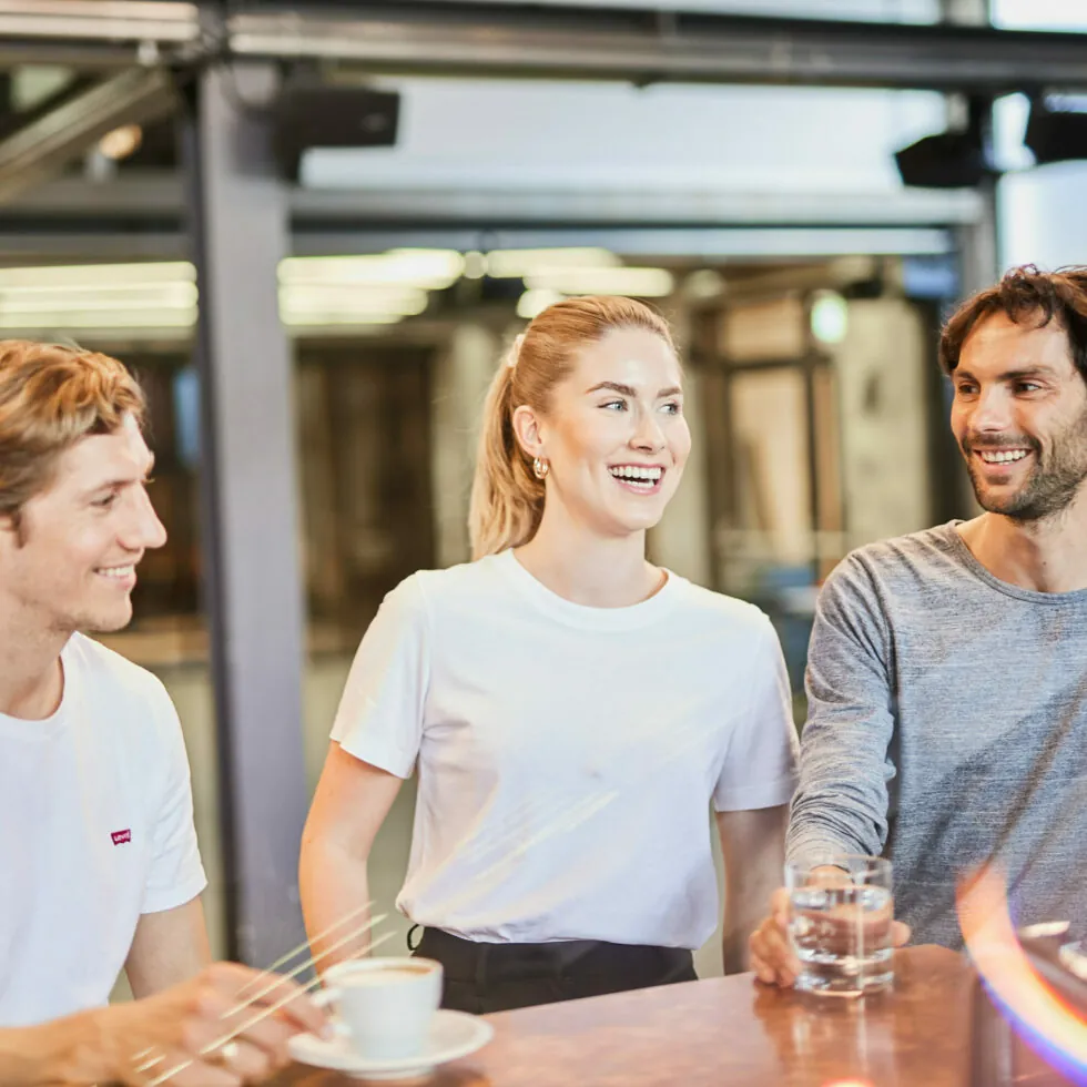 Three people are sitting at a table, smiling, and chatting. One person holds a cup, the second person is laughing, and the third person holds a glass of water. They are casually dressed.