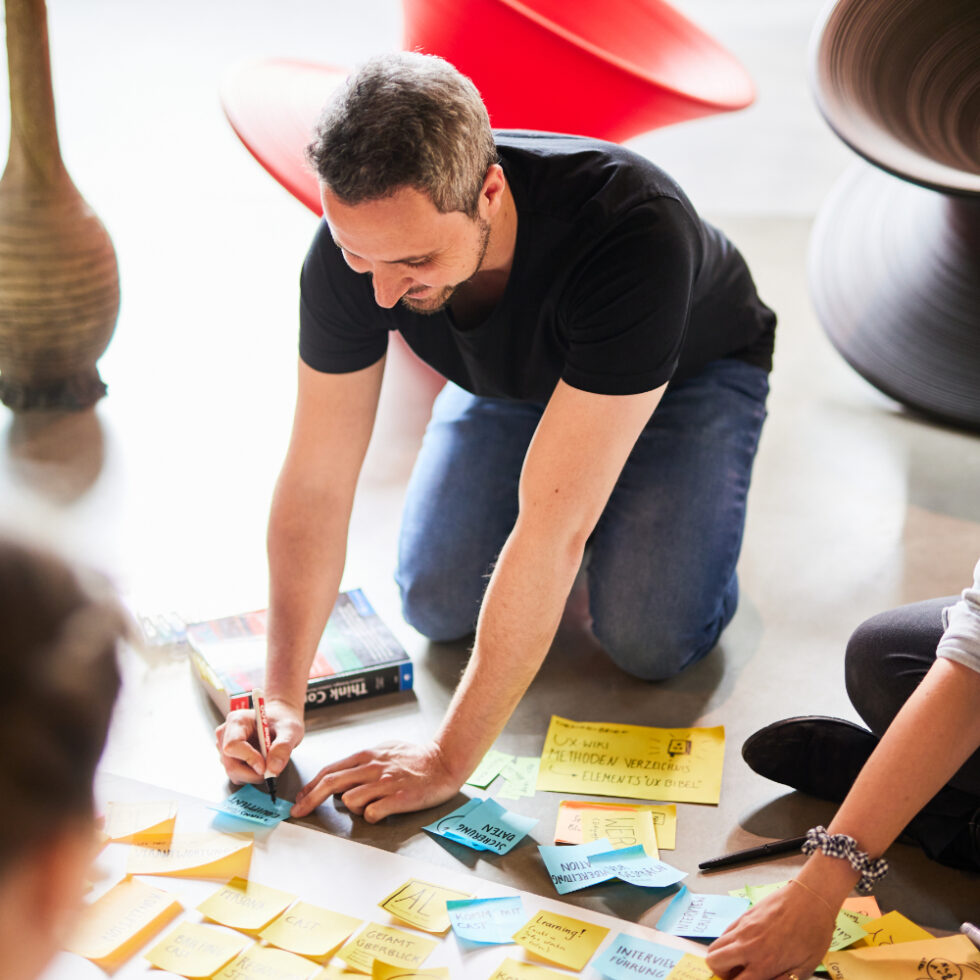 A man kneels on the floor in a workshop and writes notes on sticky notes spread out around him.