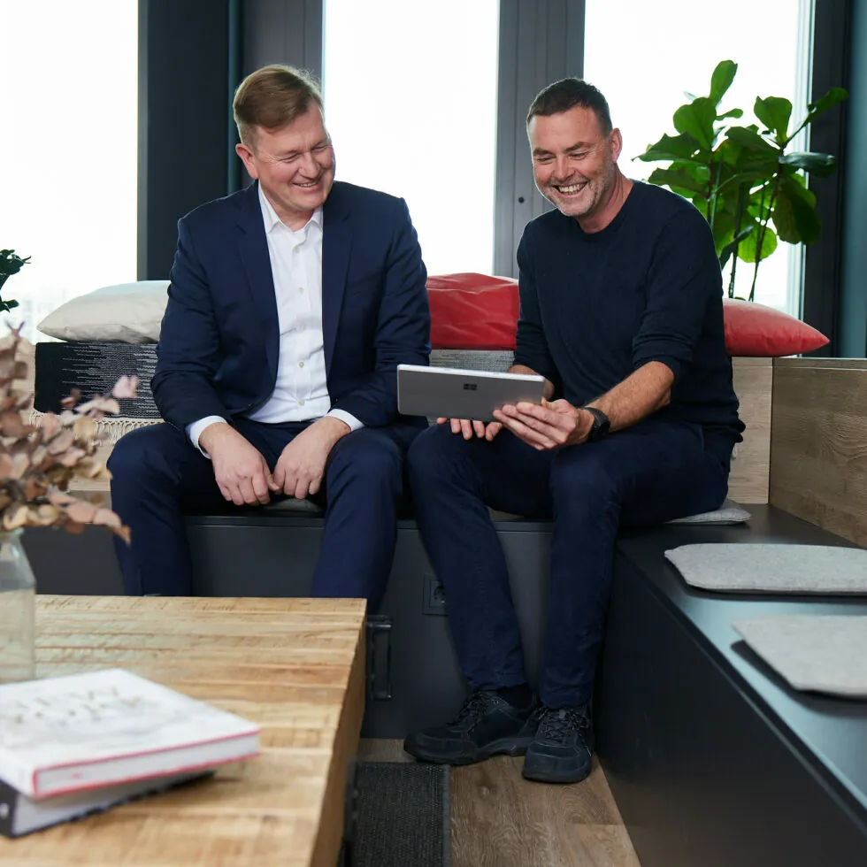 Two men sit in a modern office space. One in a suit, the other in casual wear, both smiling and looking at a tablet. A wooden table with books and a vase is in front of them.