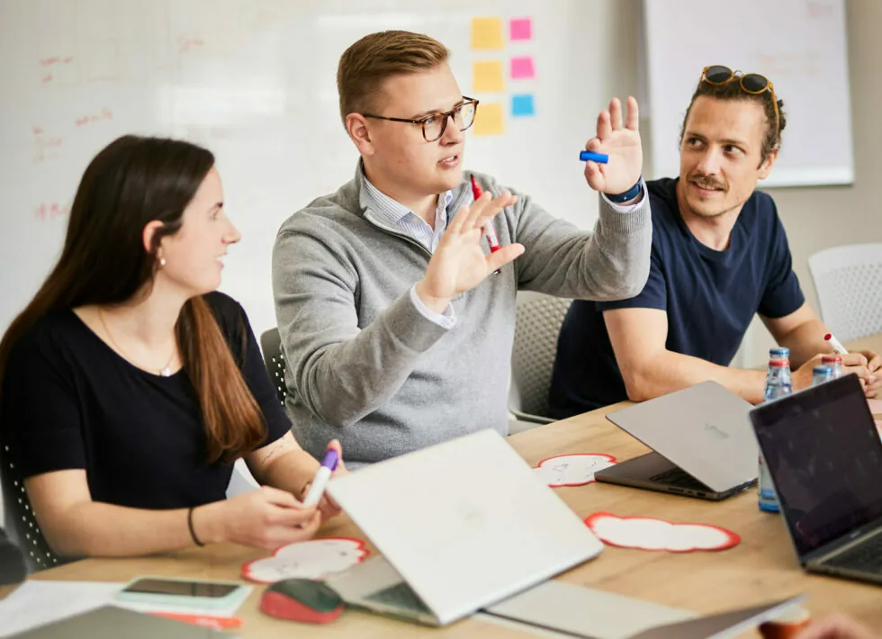 Three people having a discussion around a table with laptops and notebooks in a meeting room. One person is gesturing with a marker, while the others listen.