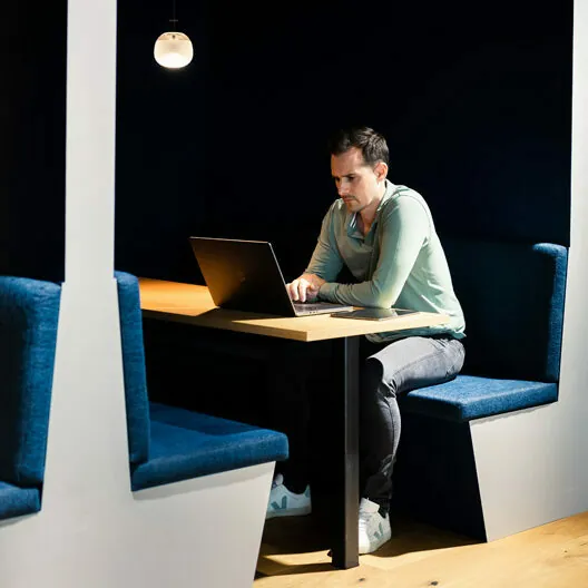 A person working on a laptop while seated in a small, modern booth with blue cushioned seats, illuminated by a single hanging light.