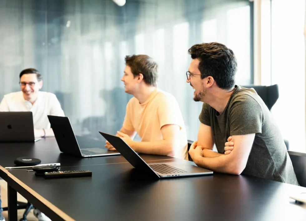 Three men sit at a conference table with laptops, engaged in conversation. A remote control and a wireless mouse are on the table.