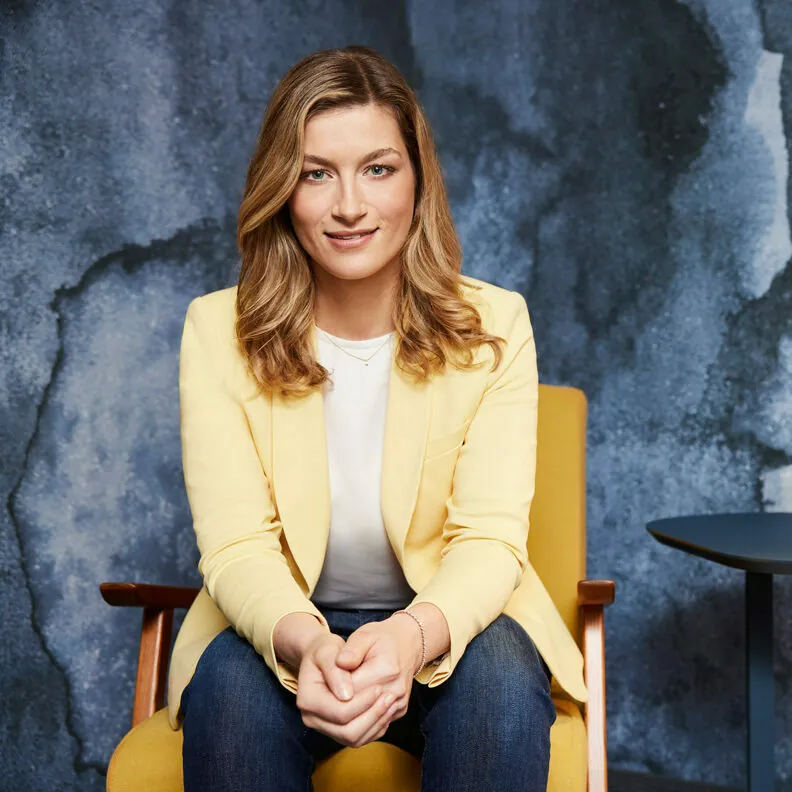 A woman with long blonde hair sits in a yellow chair, wearing a yellow blazer over a white shirt and blue jeans, against a blue abstract background.