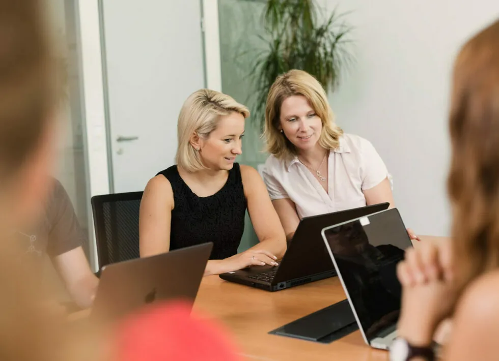 Two women are seated at a desk, working on laptops. One woman points at the screen while the other looks on. Two other individuals are also present, with only parts of their arms and shoulders visible.