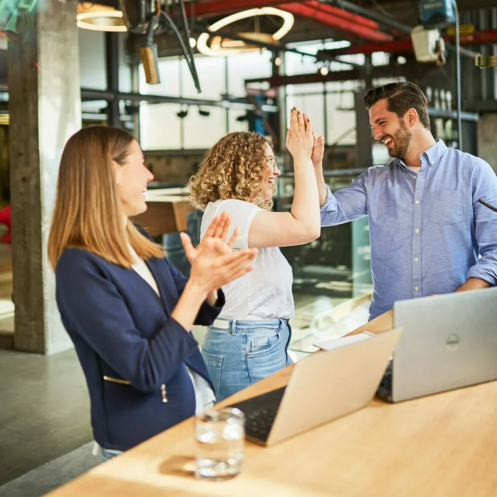 Three people celebrate a success at a workplace by high-fiving and clapping near a table with laptops and a glass of water. The office setting includes modern decor and large windows.