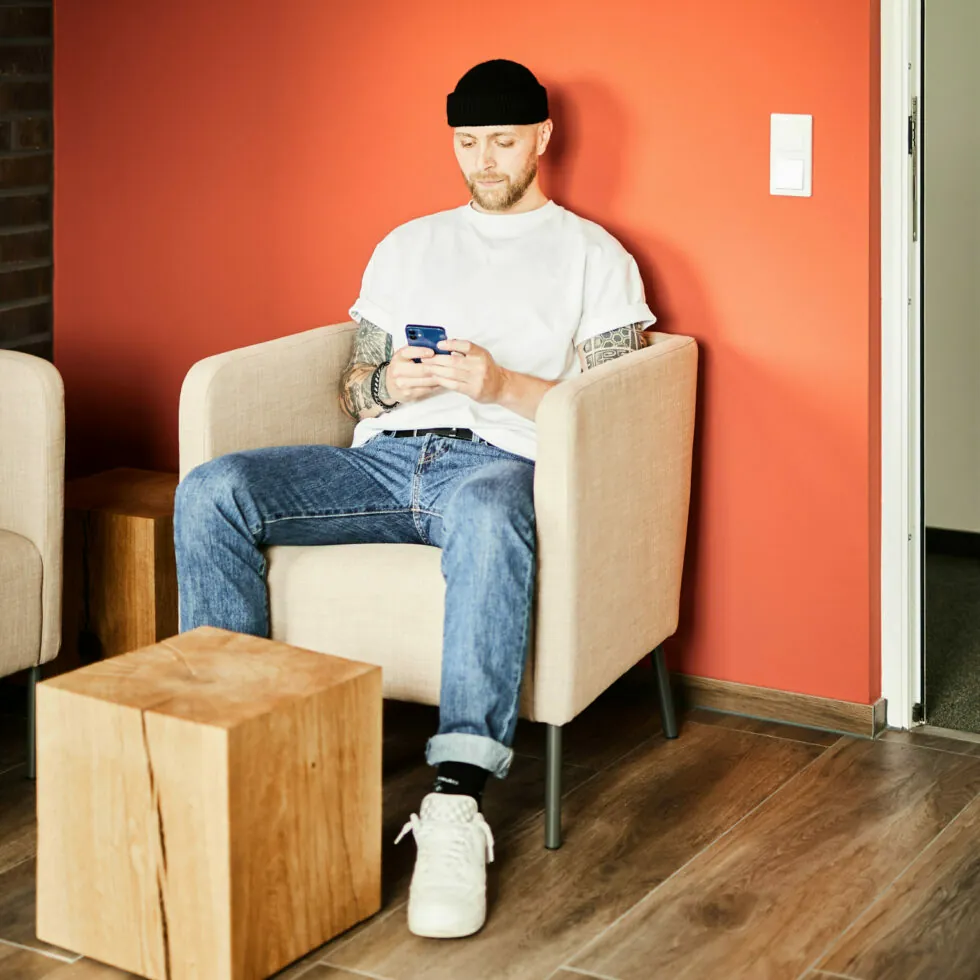 A person wearing a black beanie, white t-shirt, and jeans sits on a beige chair in front of an orange wall, looking at a phone.