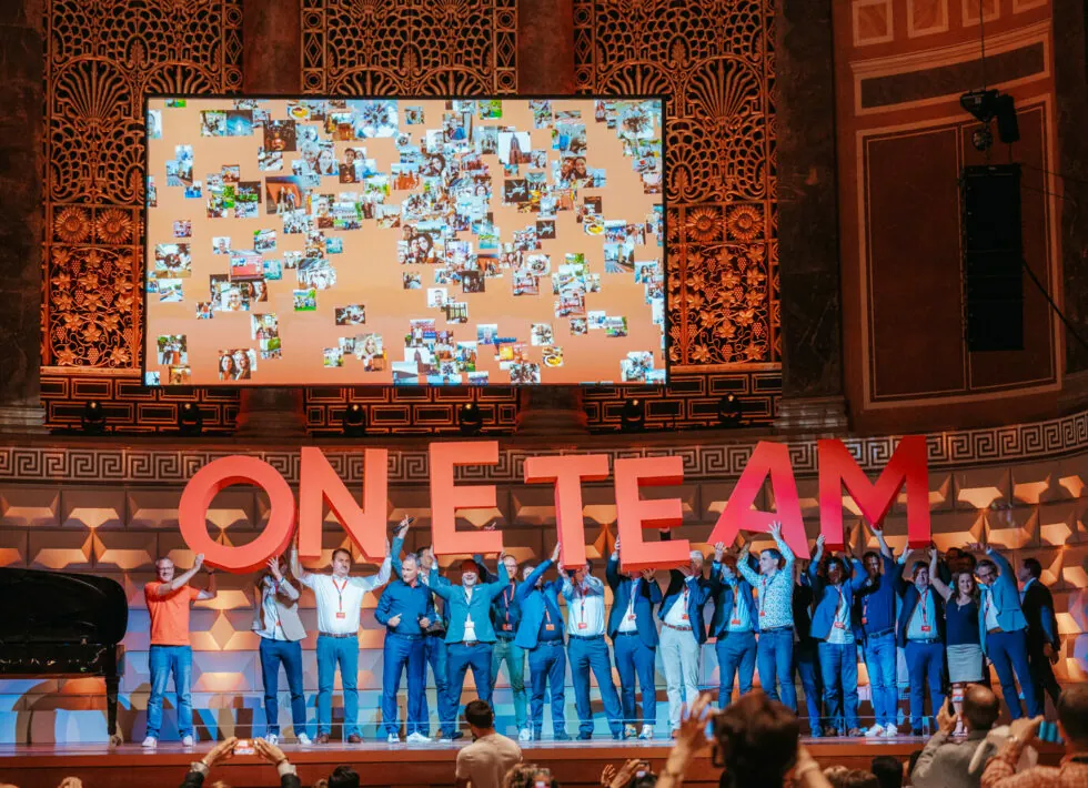 A group of people on stage holding large letters spelling "ONE TEAM" with a large screen displaying various images in the background. The event appears to be held in an ornate hall.