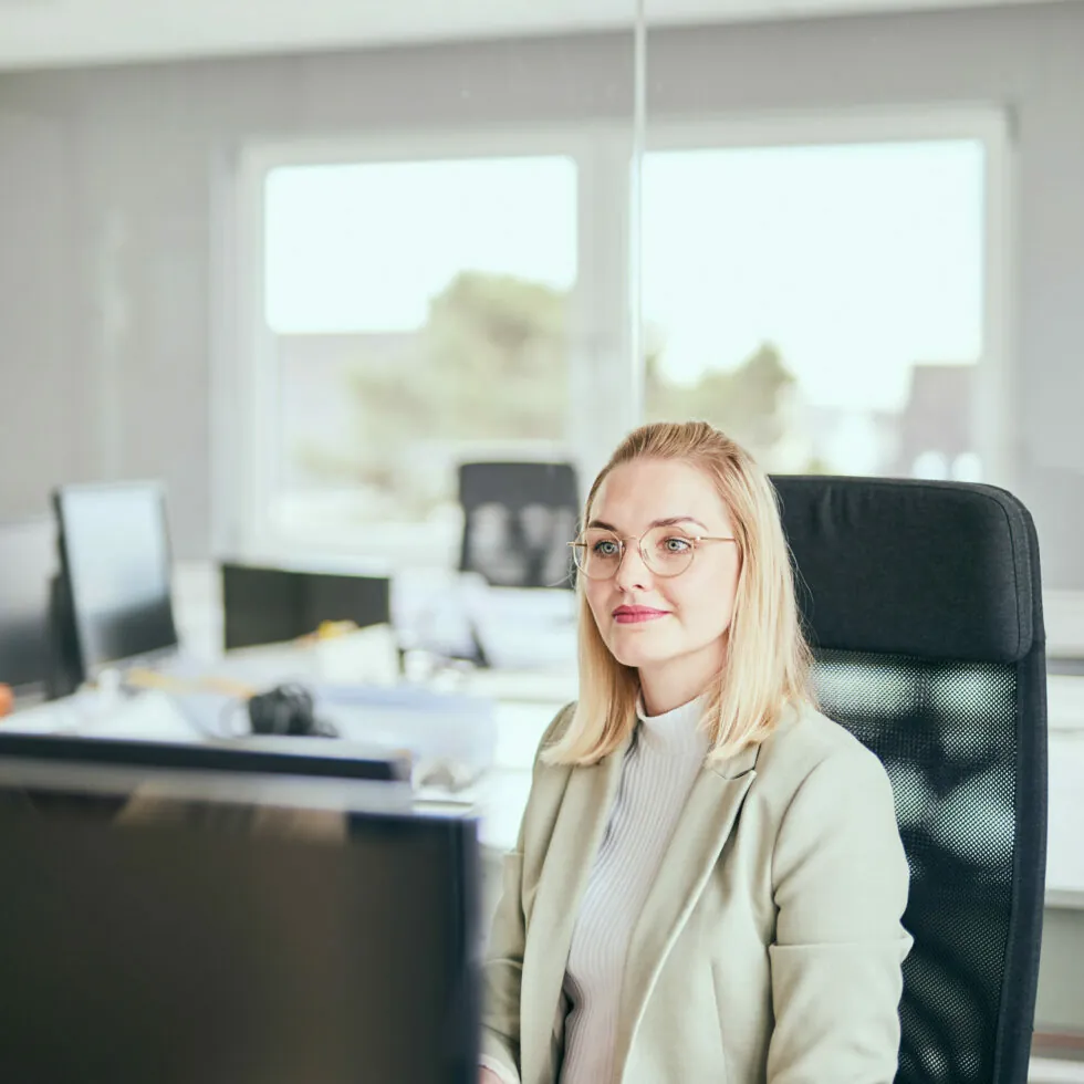 A woman with glasses and blonde hair sits at a desk with a computer in a modern office, looking ahead.
