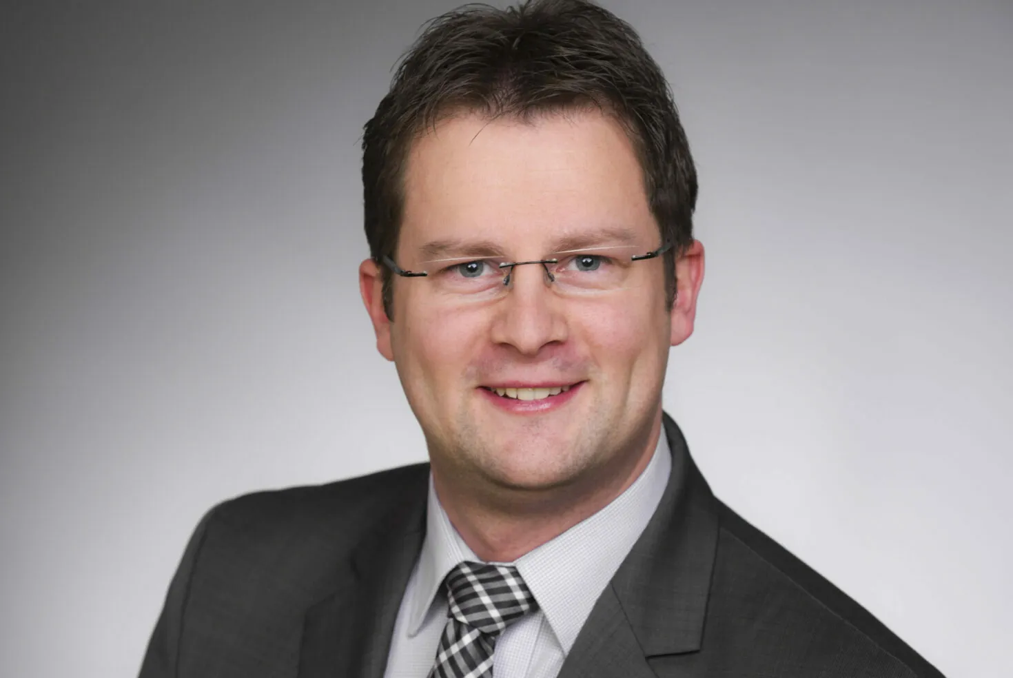 A man in a suit and striped tie is smiling at the camera. He has short dark hair, glasses, and is posing against a plain grey background.