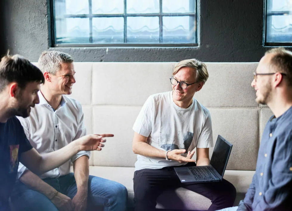 Four men are sitting on a couch, engaged in conversation and smiling. One man is holding a laptop. They are in a room with dark walls and large windows.