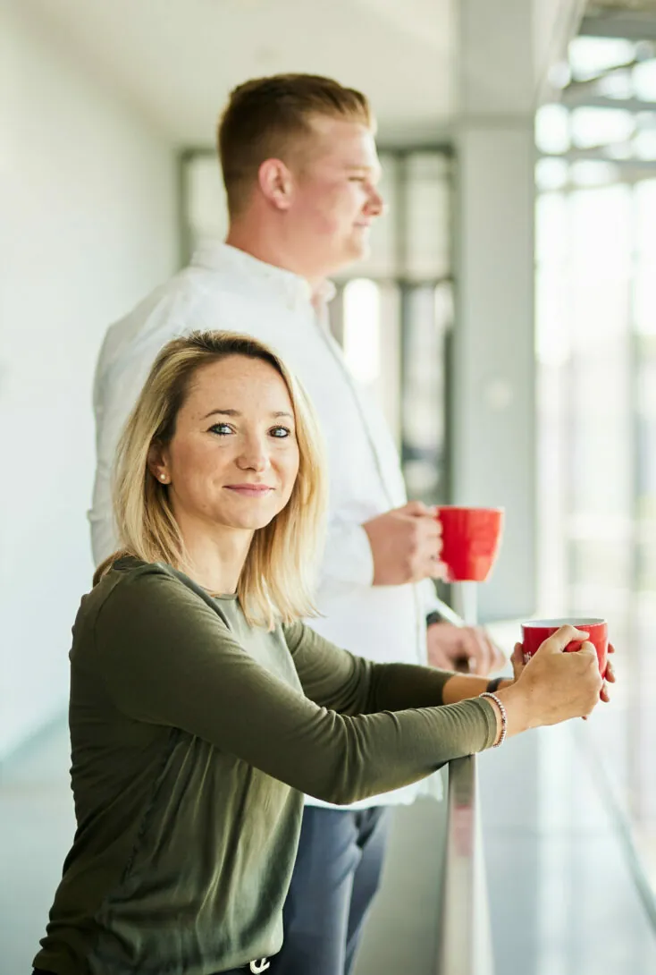 Two people are standing beside a railing, each holding a red coffee mug. One person is facing sideways, and the other is looking at the camera and smiling.
