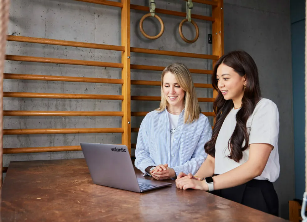 Two women stand at a wooden table, looking at a laptop. Wooden gymnastic rings and wall bars are visible in the background.