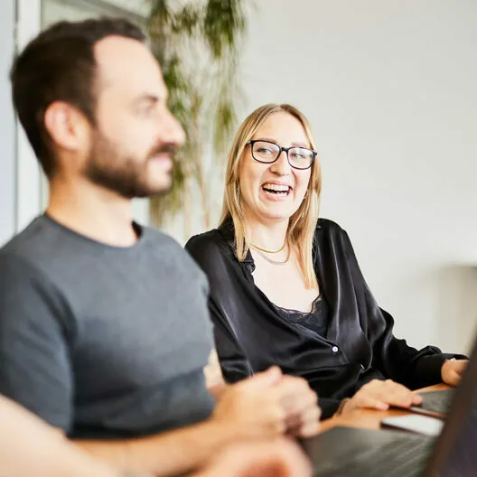 A woman smiles and talks while seated next to a man at a table with laptops.