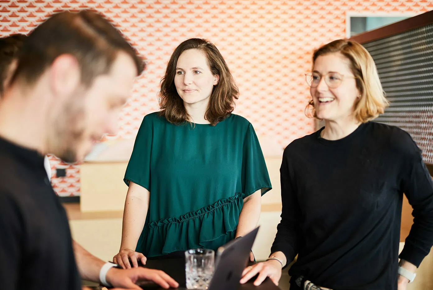 Three individuals are standing in an office, with two women smiling and a man looking at a laptop. The background features a patterned wall.