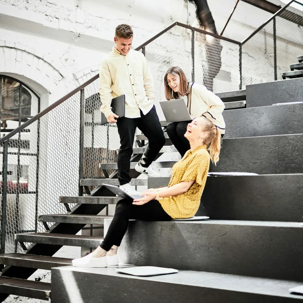 Three people with laptops are sitting and standing on industrial-style staircase steps, conversing and smiling in a bright, modern setting.