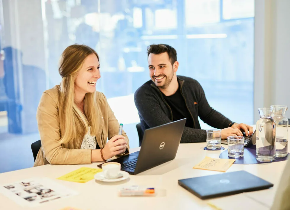 Two people sit at a desk with a laptop, water pitcher, and documents, smiling and having a conversation.