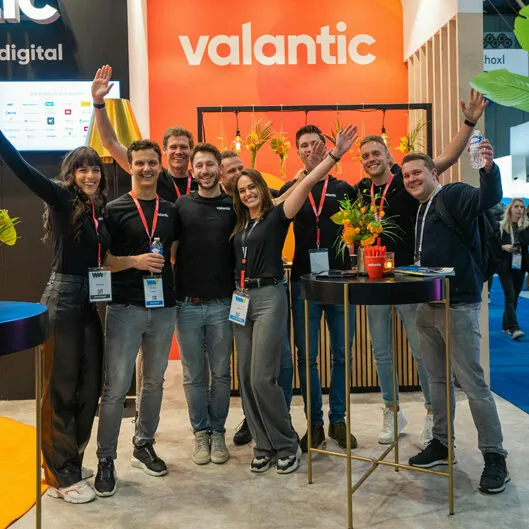 A group of eight people wearing name tags and casual clothing poses for a photo in front of an orange "valantic" booth at a trade show. Some have their arms raised in celebration.