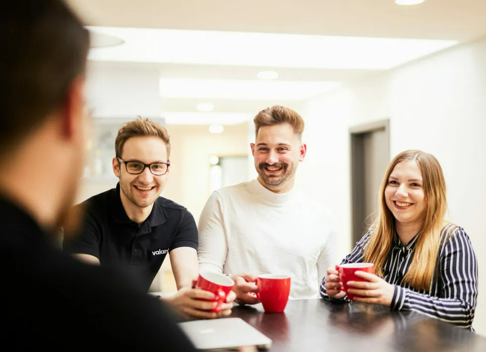 Three people sit and smile while holding red mugs, engaged in conversation with a fourth person whose back is to the camera.