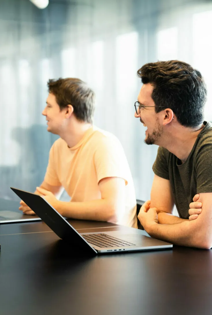 Two men sitting at a table with laptops, engaged in a discussion. The man in the foreground is laughing, while the man next to him is looking in the same direction, smiling.