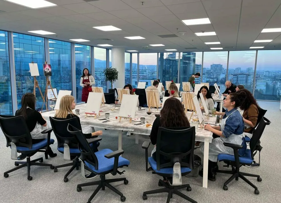 A group of people participates in a painting class inside a modern office setting. They sit at tables with canvases and art supplies, while an instructor stands at the front providing guidance.