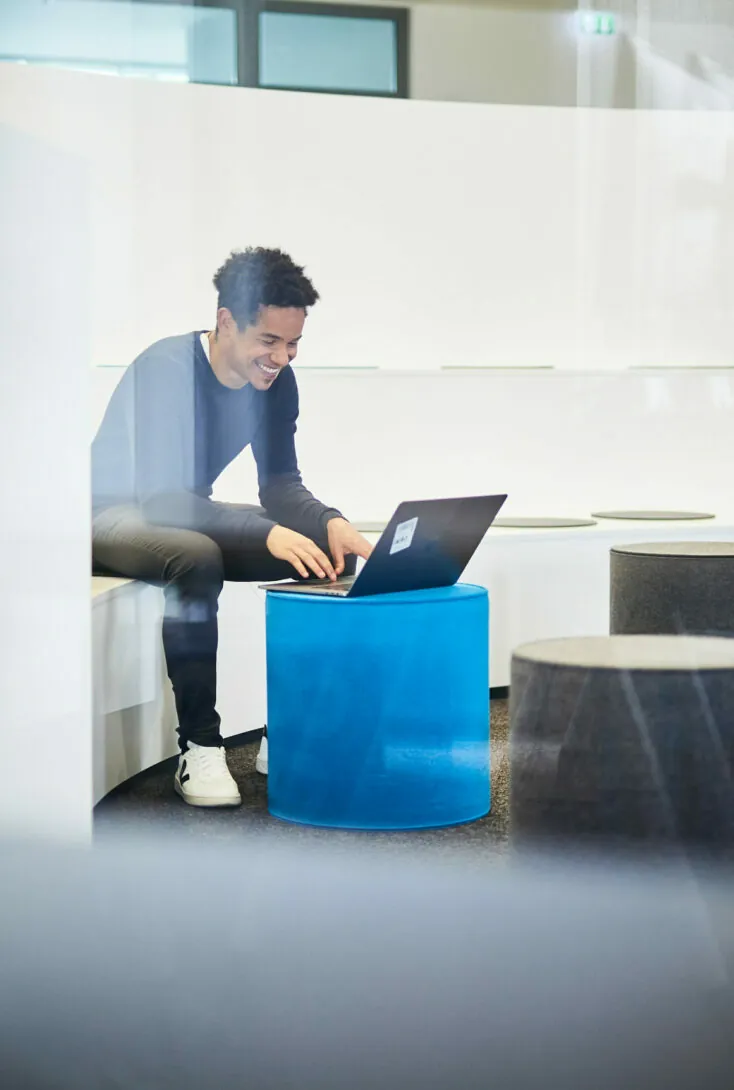 A person sits on a white bench using a laptop placed on a blue stool in a modern, minimalistic room. The individual appears engaged with the laptop.
