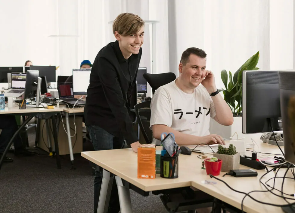 Two men are working at a desk in an office. One is seated and using a computer, while the other, standing beside him, is smiling. Various office items and decorations are visible on the desk.