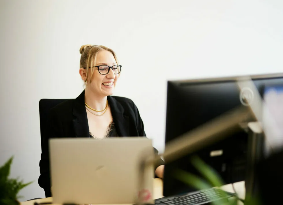A person wearing glasses and a black blazer is smiling while working at a desk with two computer monitors in an office setting.